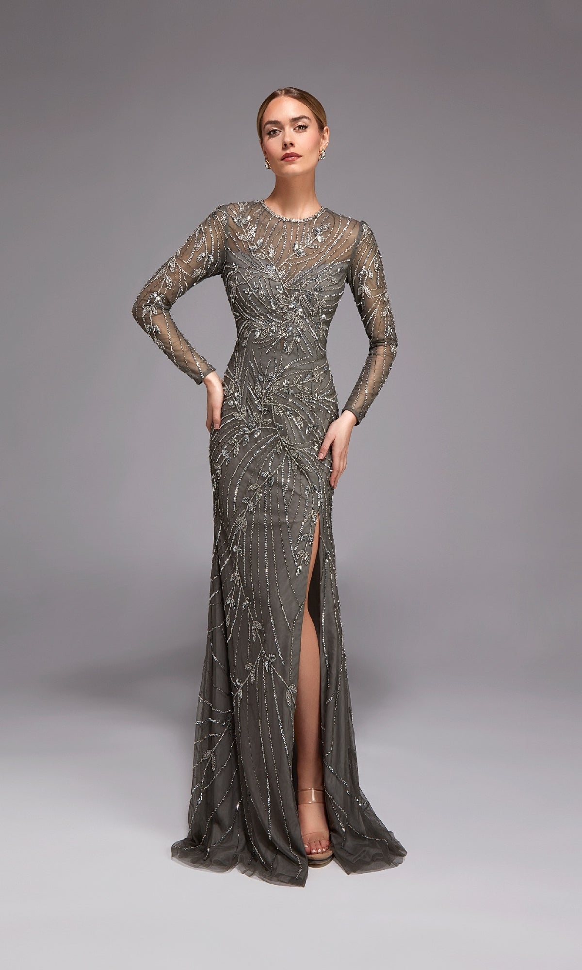 A woman poses in the Alyce 27801 Long Formal Dress—a modest gunmetal gown with silver bead embellishments, sheer long sleeves, and a thigh-high slit—set against a plain grey background.